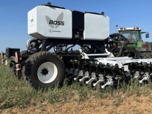 Ein Agrisem BOSS 6 HD Direktsaatgerät mit großem weißen Saatgutbehälter, angehängt an einen grünen Traktor, arbeitet auf einem trockenen Feld mit niedrigem Pflanzenbewuchs. Die Schare des Geräts sind gut sichtbar und bereit zur präzisen Saatablage.