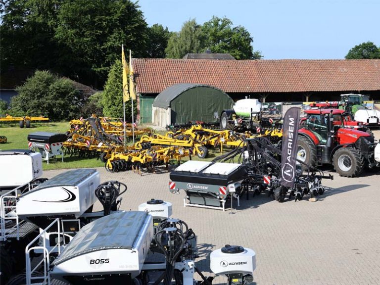 Außenaufnahme eines Hofgeländes mit landwirtschaftlichen Maschinen von AGRISEM. Im Vordergrund verschiedene Tanks und Maschinen in Weiß und Schwarz mit AGRISEM-Logo, darunter BOSS-Tanks. Im Hintergrund mehrere gelbe Bodenbearbeitungsgeräte, ein Traktor, ein schwarzer AGRISEM-Werbeflagge, sowie Scheunen und grüne Bäume.