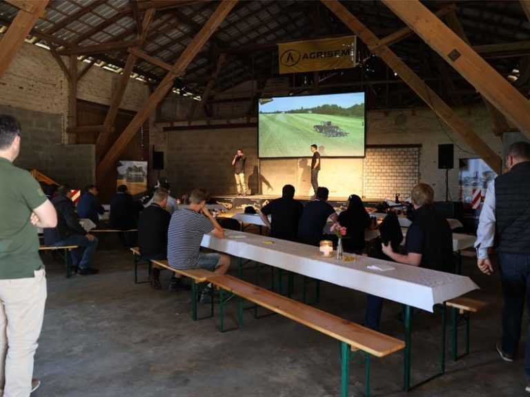 Innenaufnahme einer Scheune mit Holzbalken, in der eine AGRISEM Veranstaltung stattfindet. Menschen sitzen auf Bierbänken an langen Tischen mit weißen Tischdecken. Im Hintergrund steht eine Bühne mit zwei Personen und einem großen Bildschirm, der ein Bild von einem Traktor mit AGRISEM Maschine auf dem Feld zeigt. Über der Bühne hängt ein AGRISEM-Banner.