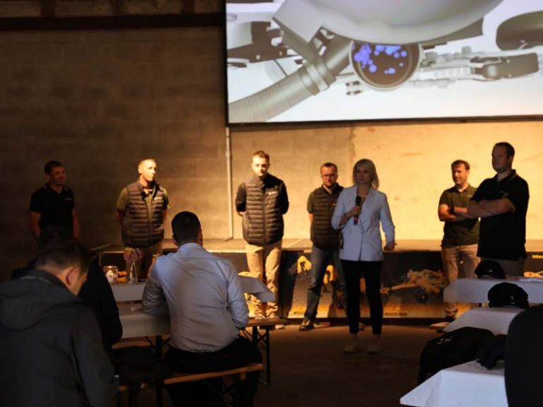 Innenaufnahme einer Scheune mit einer AGRISEM Veranstaltung. Im Vordergrund sitzen Teilnehmer an Biertischen mit weißen Tischdecken. Im Hintergrund stehen sieben Personen, darunter eine Frau mit Mikrofon, vor einer Bühne. Hinter ihnen ist eine Leinwand mit einer technischen Illustration eines AGRISEM Geräts zu sehen.