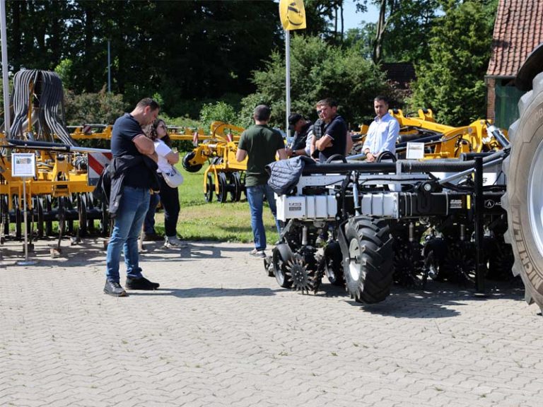 Menschen bei einer AGRISEM Außenausstellung auf einem Hof. Im Vordergrund ein landwirtschaftliches Gerät mit schwarzen Scheiben und Schläuchen, daneben ein Traktor. Mehrere Besucher stehen in Gruppen und betrachten die ausgestellten Maschinen. Im Hintergrund sind weitere gelbe AGRISEM Anbaugeräte und Bäume zu sehen.