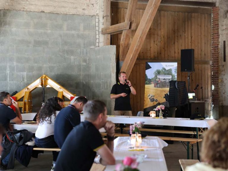 Lars-Wilhelm Funk steht in einer Scheune und hält mit einem Mikrofon einen Vortrag vor sitzenden Gästen an Biertischen. Er trägt ein schwarzes AGRISEM-Polo. Im Hintergrund stehen ein gelbes landwirtschaftliches Gerät und ein Roll-Up-Banner mit einem Bild von AGRISEM-Maschinen. Auf den Tischen stehen Blumenvasen und Kerzen.