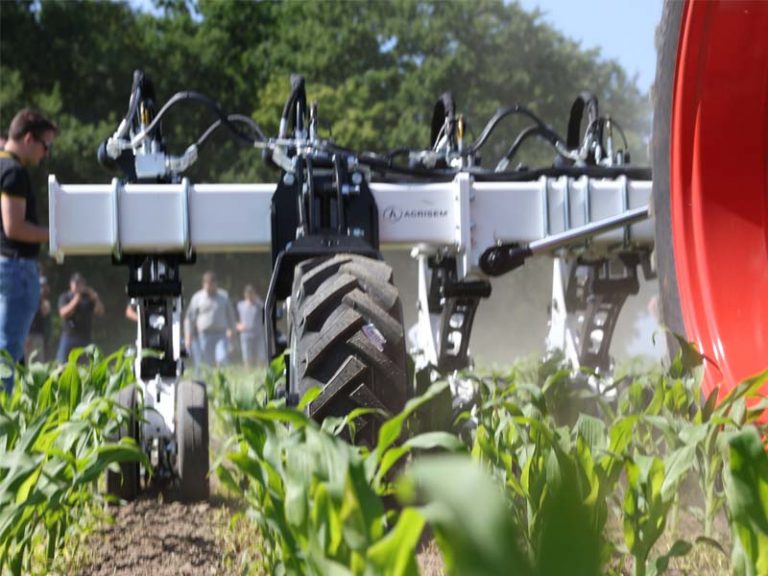 Nahaufnahme der AGRISEM SCALP Maschine während einer Feldvorführung. Das Gerät arbeitet zwischen den Pflanzenreihen auf einem Feld mit jungem Mais. Im Vordergrund ist das große Traktorrad mit roter Felge sichtbar, im Hintergrund Zuschauer und Bäume.