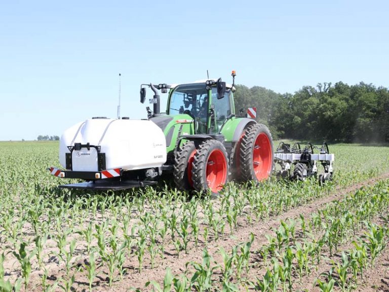 Grüner Traktor mit Doppelbereifung auf dem Acker bei sonnigem Wetter. Am Frontanbau ist ein weißer Fronttank vom Typ Agrisem DSL montiert, am Heck arbeitet eine Agrisem Scalp Maschine zur Bodenbearbeitung zwischen den Reihen von jungen Maispflanzen.
