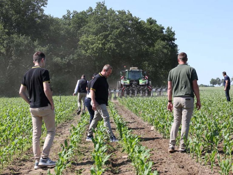 Gruppe von Menschen läuft zwischen Maisreihen auf einem Feld. Im Hintergrund fährt ein grüner Traktor mit roter Doppelbereifung und angebauter Agrisem Maschine auf die Gruppe zu. Der Boden ist leicht staubig, und im Hintergrund stehen Bäume unter blauem Himmel.