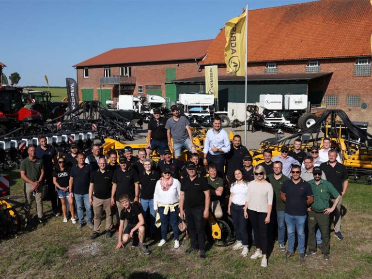 Gruppenfoto von etwa 40 Personen vor einer landwirtschaftlichen Maschinenhalle mit roten Dächern. Die Menschen, bestehend aus Männern und Frauen, tragen überwiegend schwarze Agrisem-Poloshirts oder Freizeitkleidung. Im Hintergrund stehen gelbe und weiße Agrisem-Landmaschinen, mehrere Fahnen mit Agrisem-Logo wehen im Wind. Die Gruppe posiert auf einer Wiese bei sonnigem Wetter.