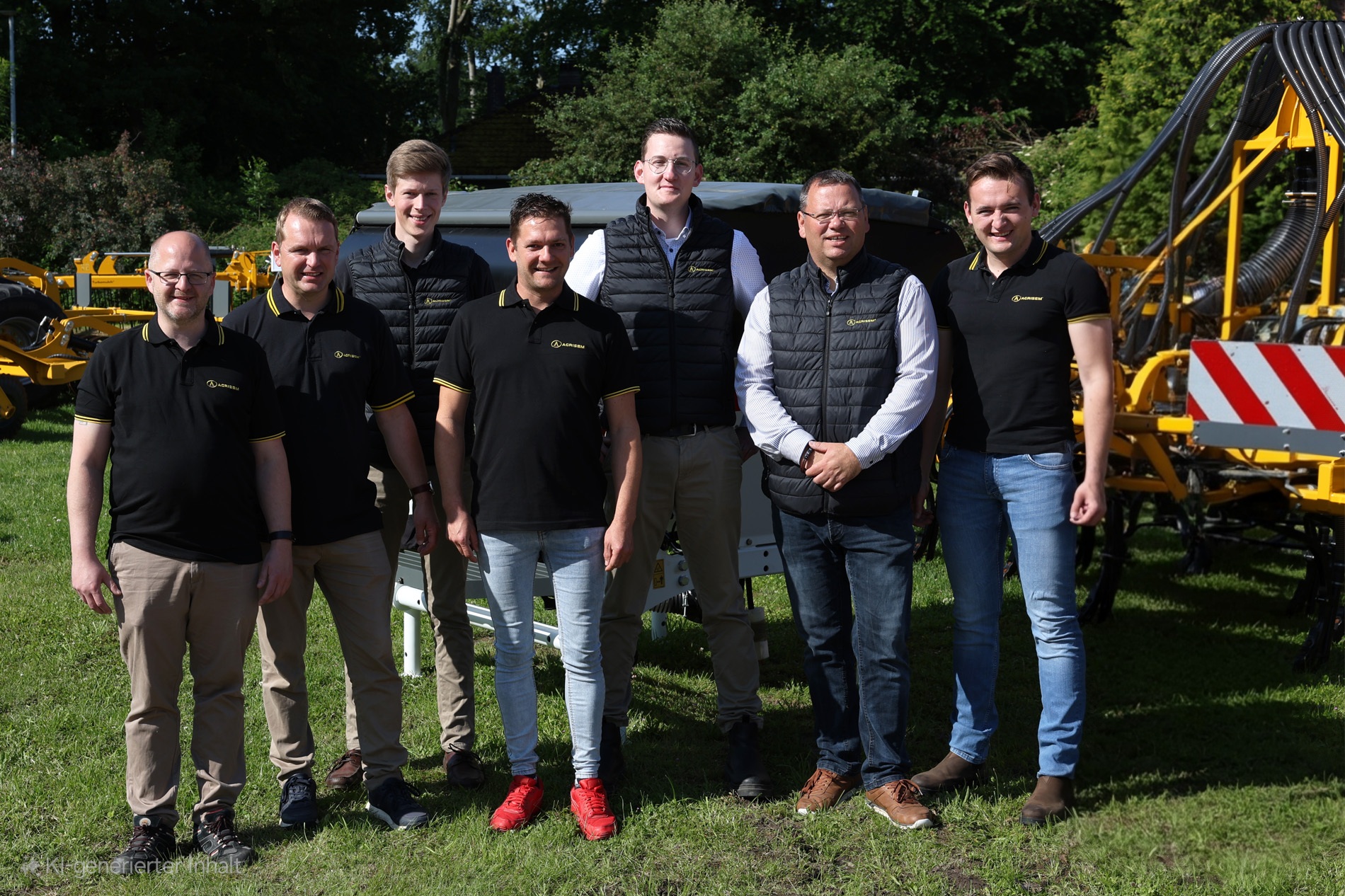 Gruppenbild des AGRISEM Deutschland Teams im Freien vor gelben landwirtschaftlichen Maschinen. Sieben Männer stehen nebeneinander auf einer grünen Wiese, einige tragen schwarze AGRISEM-Poloshirts mit gelber Paspelierung, andere schwarze Westen mit AGRISEM-Logo. Im Hintergrund sind Maschinen und Bäume zu sehen.