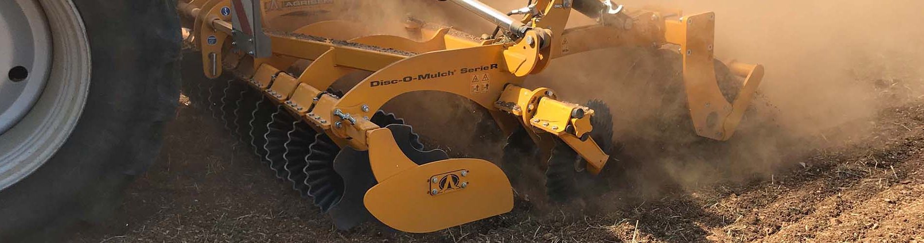 Ein Agrisem Disc-O-Mulch R arbeitet auf einem Feld hinter einem Traktor und erzeugt eine große Staubwolke. Die gelbe Scheibenegge ist mit mehreren gezackten Scheiben ausgestattet, die den Boden zerkleinern und mischen. Der Boden wird gleichmäßig bearbeitet, während das hintere Packersystem für eine Rückverfestigung sorgt. Die Hydraulikzylinder sind sichtbar, um die Arbeitstiefe anzupassen.