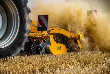 Ein Agrisem Disc-O-Mulch R arbeitet auf einem Stoppelfeld und wirbelt eine große Staub- und Erdwolke auf. Die gezackten Scheiben der gelben Maschine durchschneiden die Ernterückstände und mischen die oberste Bodenschicht. Die rot-weißen Warntafeln an der Maschine sorgen für Sicherheit im Straßenverkehr. Der Traktor, von dem nur das große Hinterrad sichtbar ist, zieht die Scheibenegge durch das trockene Feld und hinterlässt eine gleichmäßig bearbeitete Bodenstruktur.