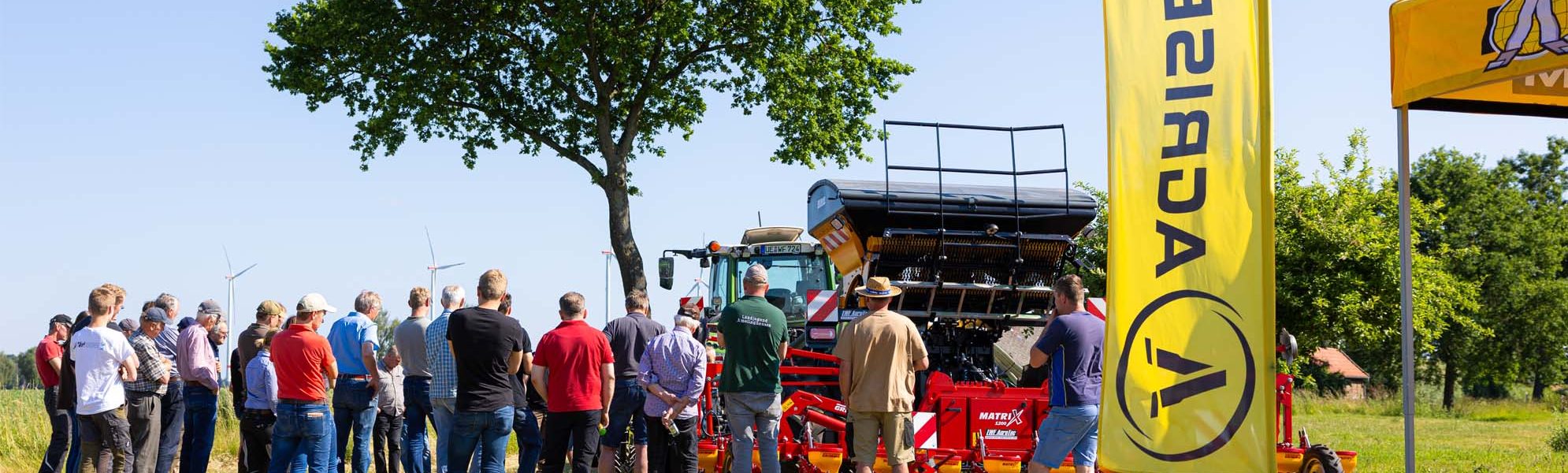 Teilnehmer eines Agrisem Deutschland Feldtages stehen auf einer Wiese und verfolgen eine Maschinenvorführung. Im Vordergrund weht eine gelbe Agrisem-Fahne, während eine Agrisem Stripcat an einem Traktor präsentiert wird.
