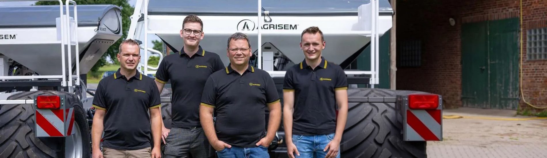 Das Team vom Vertrieb von Agrisem Deutschland steht vor zwei Agrisem-Maschinen. Die sechs Teammitglieder tragen schwarze Poloshirts mit gelben Akzenten und dem Agrisem-Logo. Sie posieren vor einer landwirtschaftlichen Kulisse mit einer Scheune im Hintergrund.